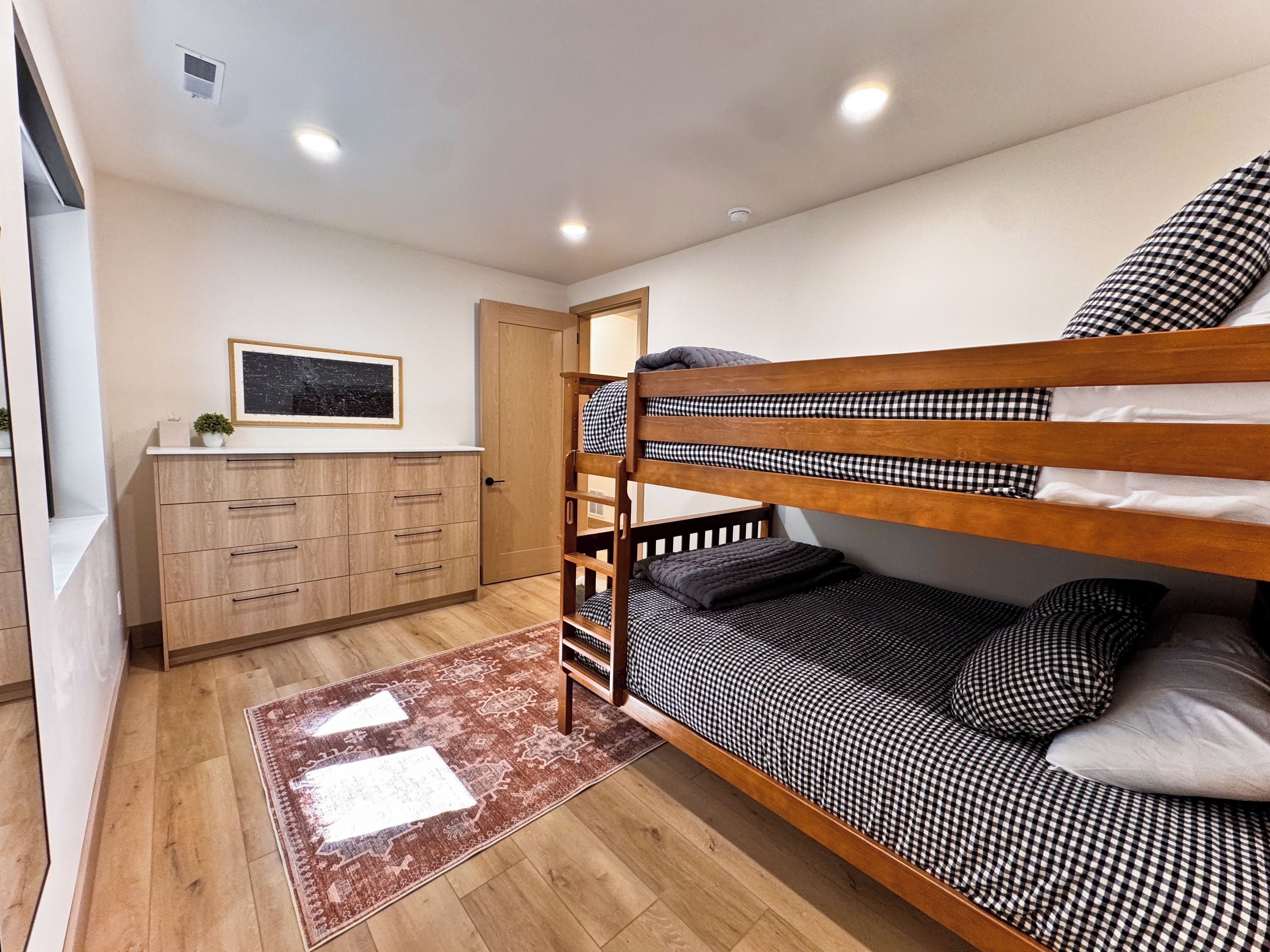 Una cama litera con ropa de cama a cuadros, un armario de madera, un cuadro en la pared y una alfombra estampada en el suelo.