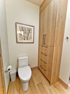 A bathroom with a toilet, a trash bin, a framed picture on the wall, and a wooden wardrobe with drawers. - 6BR Luxury Ski-In/Ski-Out Chalet with Private Sauna, Hot Tub, EV, AC (Brian Head)