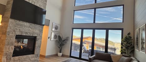 Un salon spacieux avec une cheminée en pierre, de grandes fenêtres et une vue sur l’extérieur.