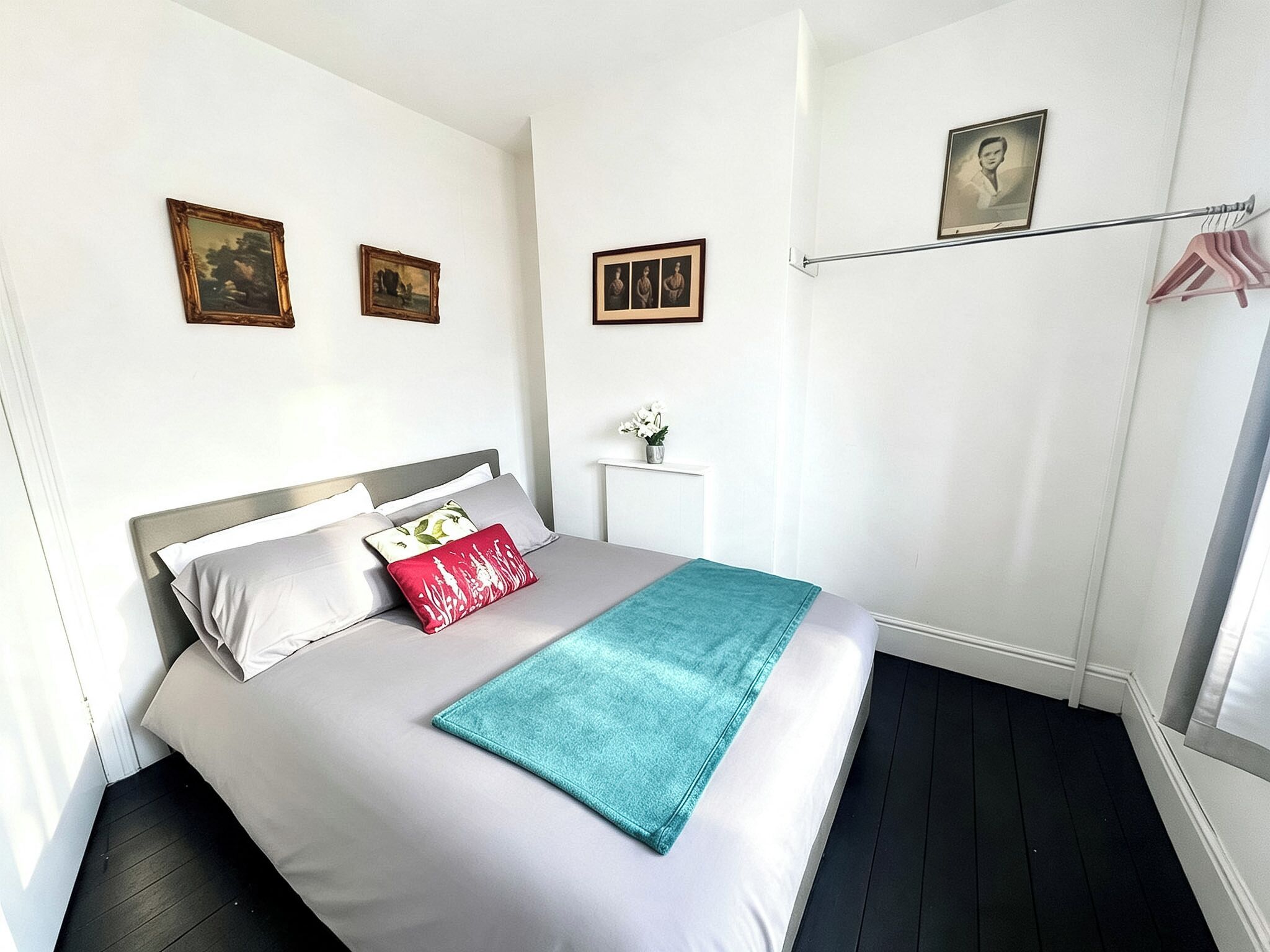 Un dormitorio ordenado con cama, almohadas, una mesita de noche con un jarrón y un cuadro colgado en la pared.