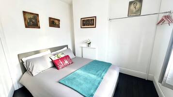 Un dormitorio ordenado con cama, almohadas, una mesita de noche con un jarrón y un cuadro colgado en la pared.