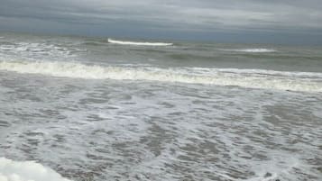 Une plage où les vagues se brisent près du rivage, un ciel nuageux et un horizon lointain.
