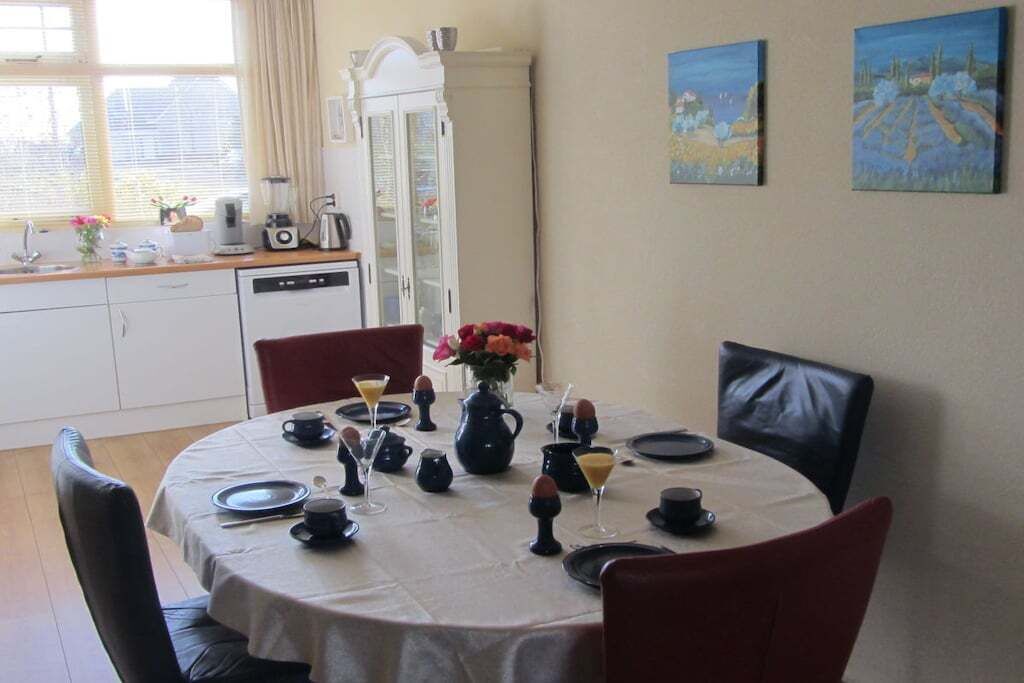 Ein Essbereich mit einem runden Tisch, gedeckt für vier Personen, umgeben von Stühlen. Im Hintergrund ist eine Küche mit weißen Schränken und einem Kühlschrank zu sehen.