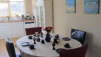 On aperçoit un coin repas avec une table ronde dressée pour quatre personnes, entourée de chaises. Au fond, on distingue une cuisine avec des meubles blancs et un réfrigérateur.