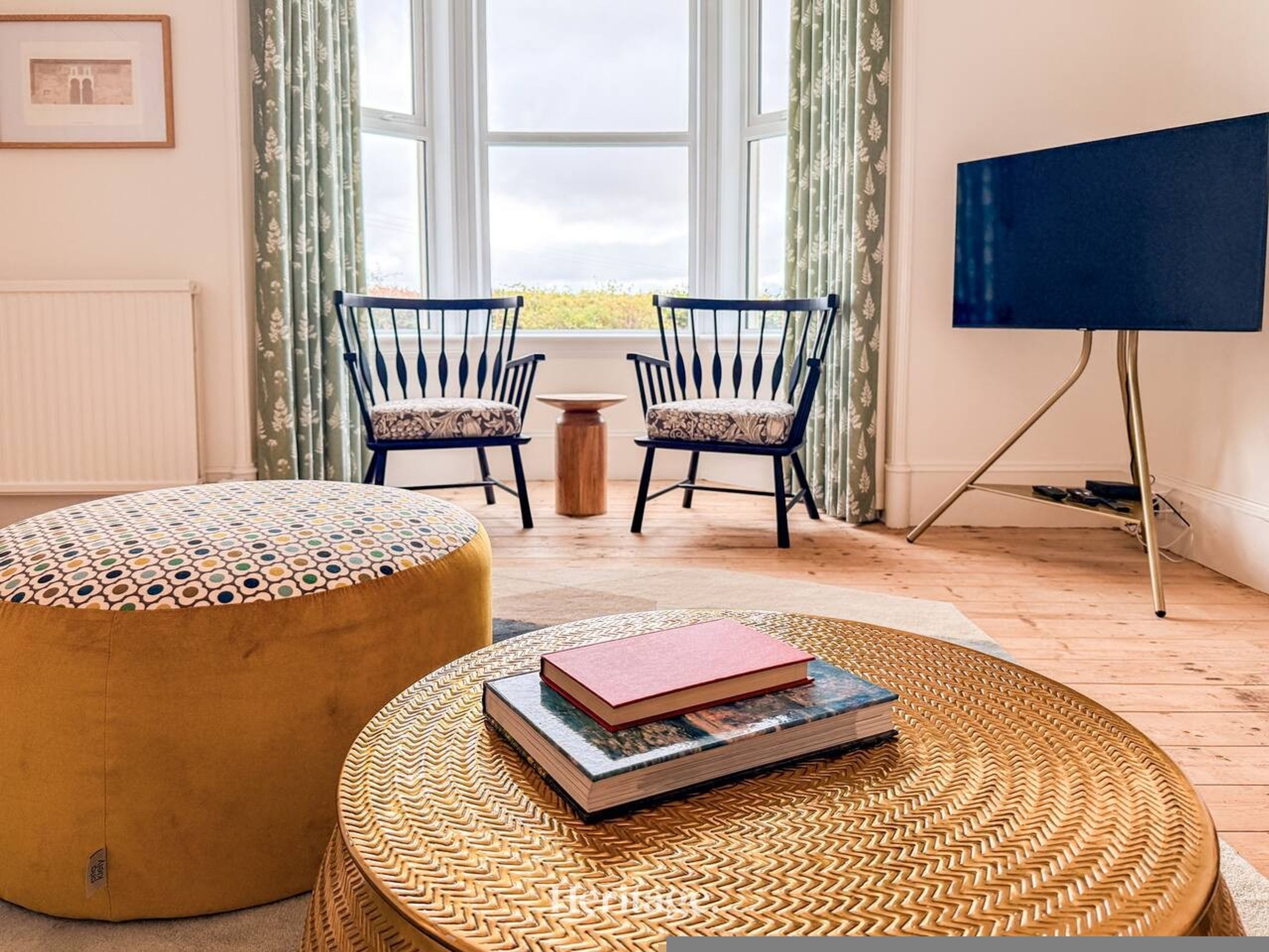 Een kamer met een groot raam, twee stoelen met gestreepte kussens, een rond poef, een kleine tafel en een televisie op een standaard.