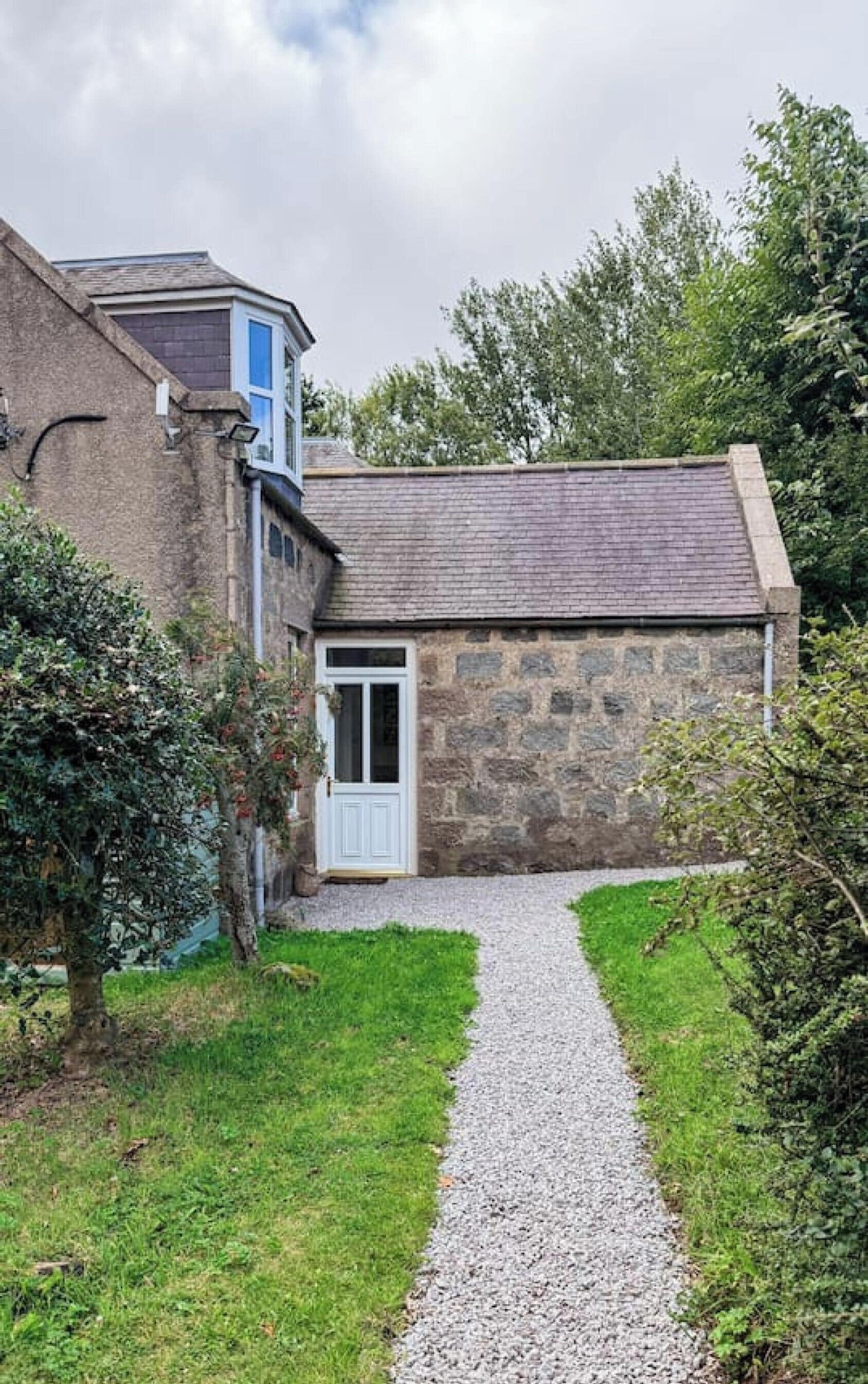 A stone building with a gravel path leading to a white door, surrounded by greenery.