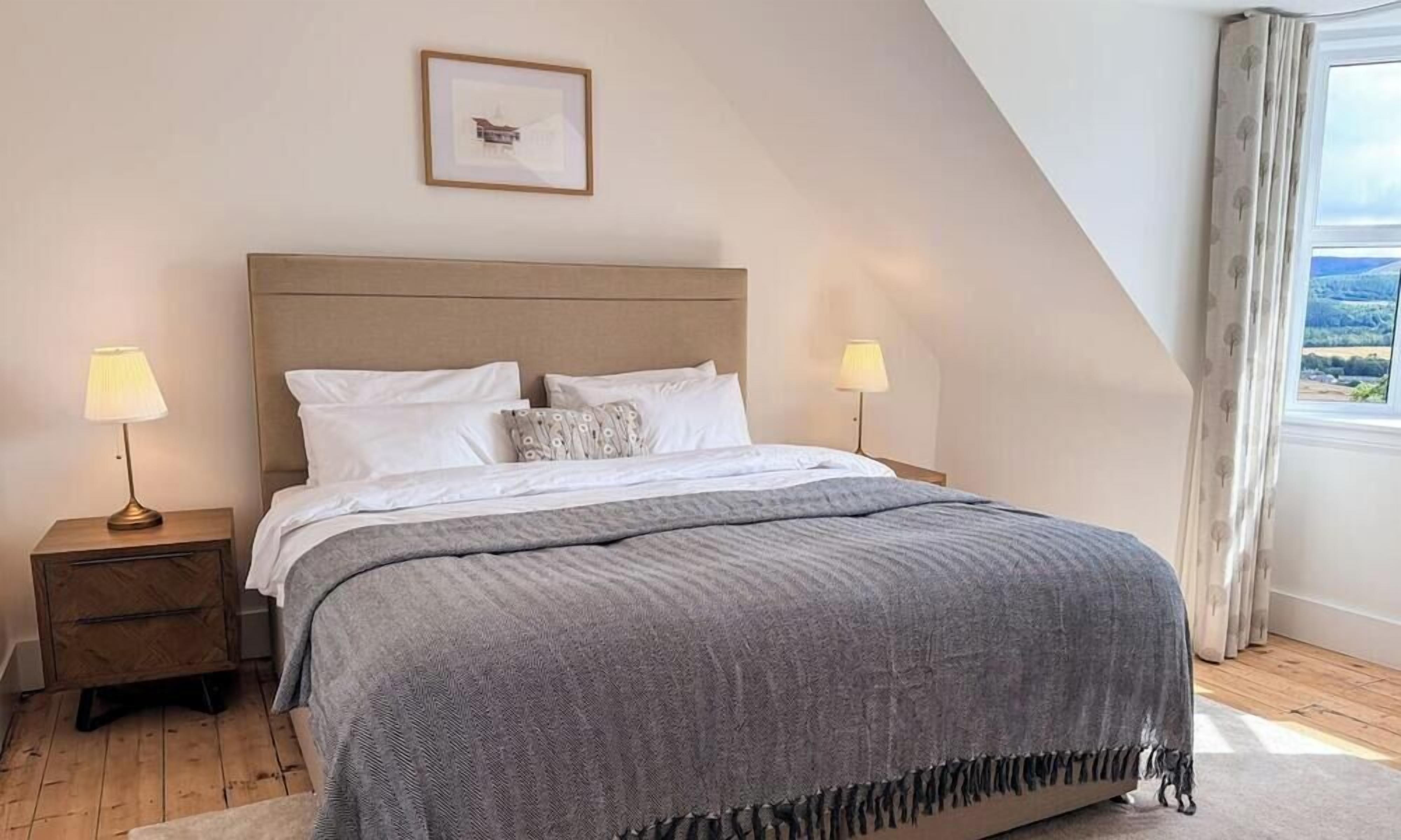 A bedroom with a large bed, two bedside tables, a framed picture on the wall, and a window with curtains.