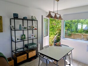 Een kamer met een eettafel, stoelen, een zwart metalen rek met diverse spullen, en een raam met uitzicht op groen.