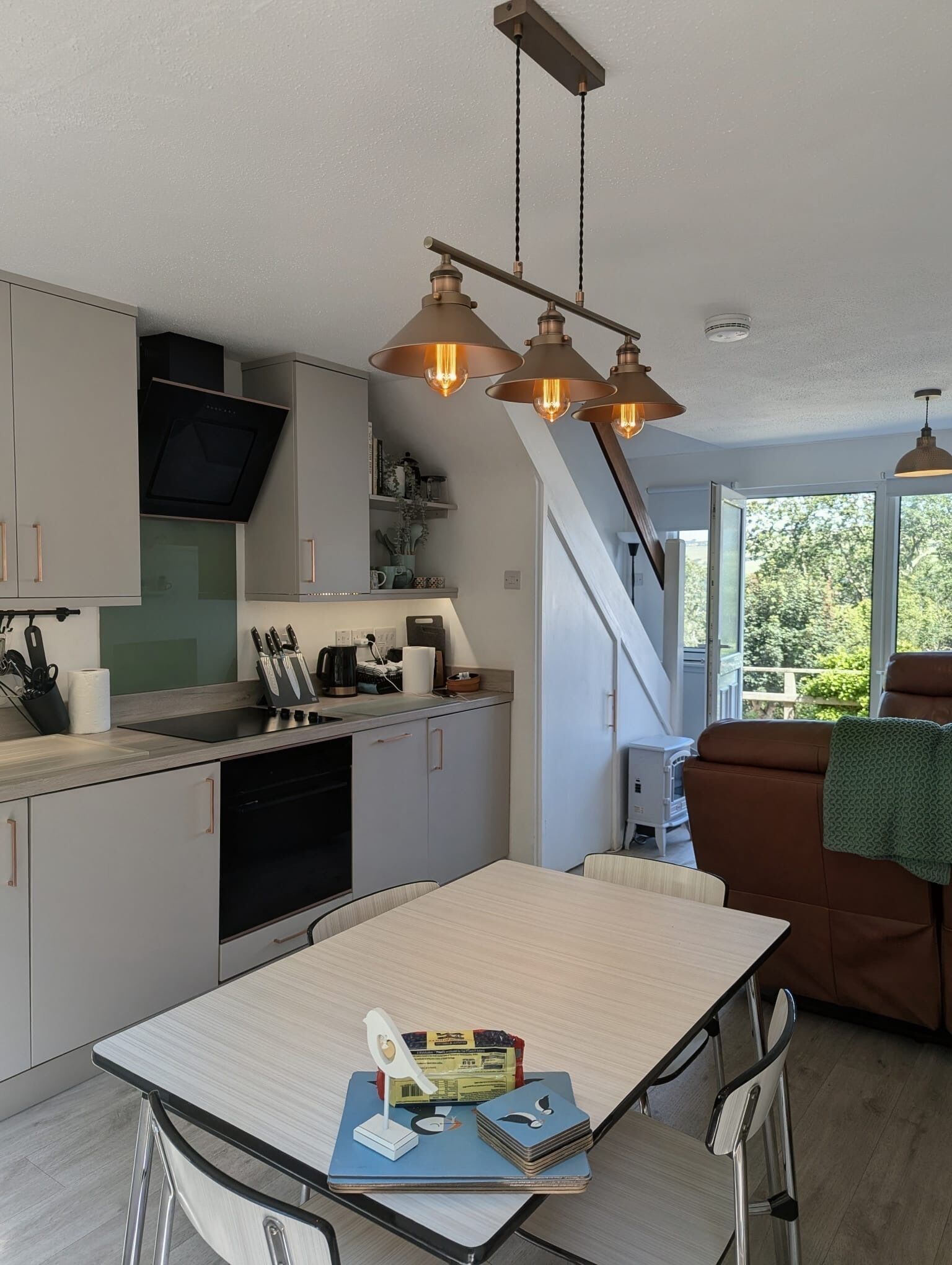 A modern kitchen with a dining area, a staircase, and a view of greenery outside.