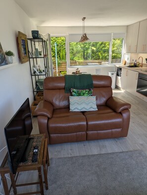 A living room with a brown leather sofa, a coffee table with a remote control, and a view of a kitchen and garden through a large window. - Sea Holly, stylish home close to lovely beach (Pembroke)