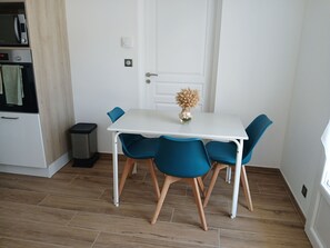 A compact dining area with a white table, four blue chairs, a small vase with dried flowers, and a trash bin. - 3-Person "Gite Étoiles" (BISCARROSSE PLAGE)