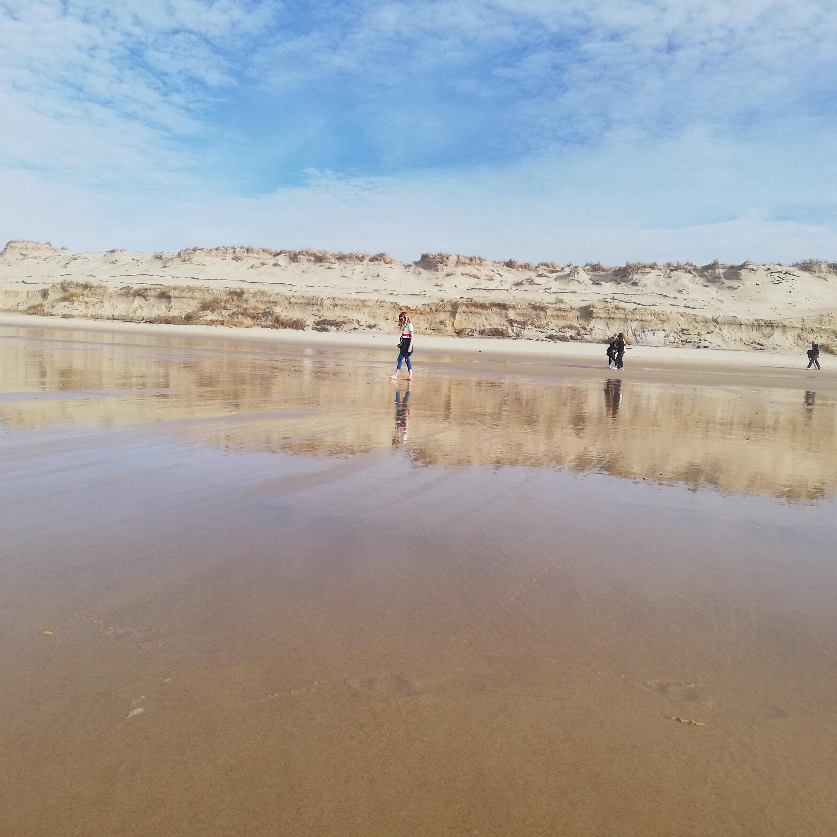 A beach with a calm water surface reflecting the sky and people walking.