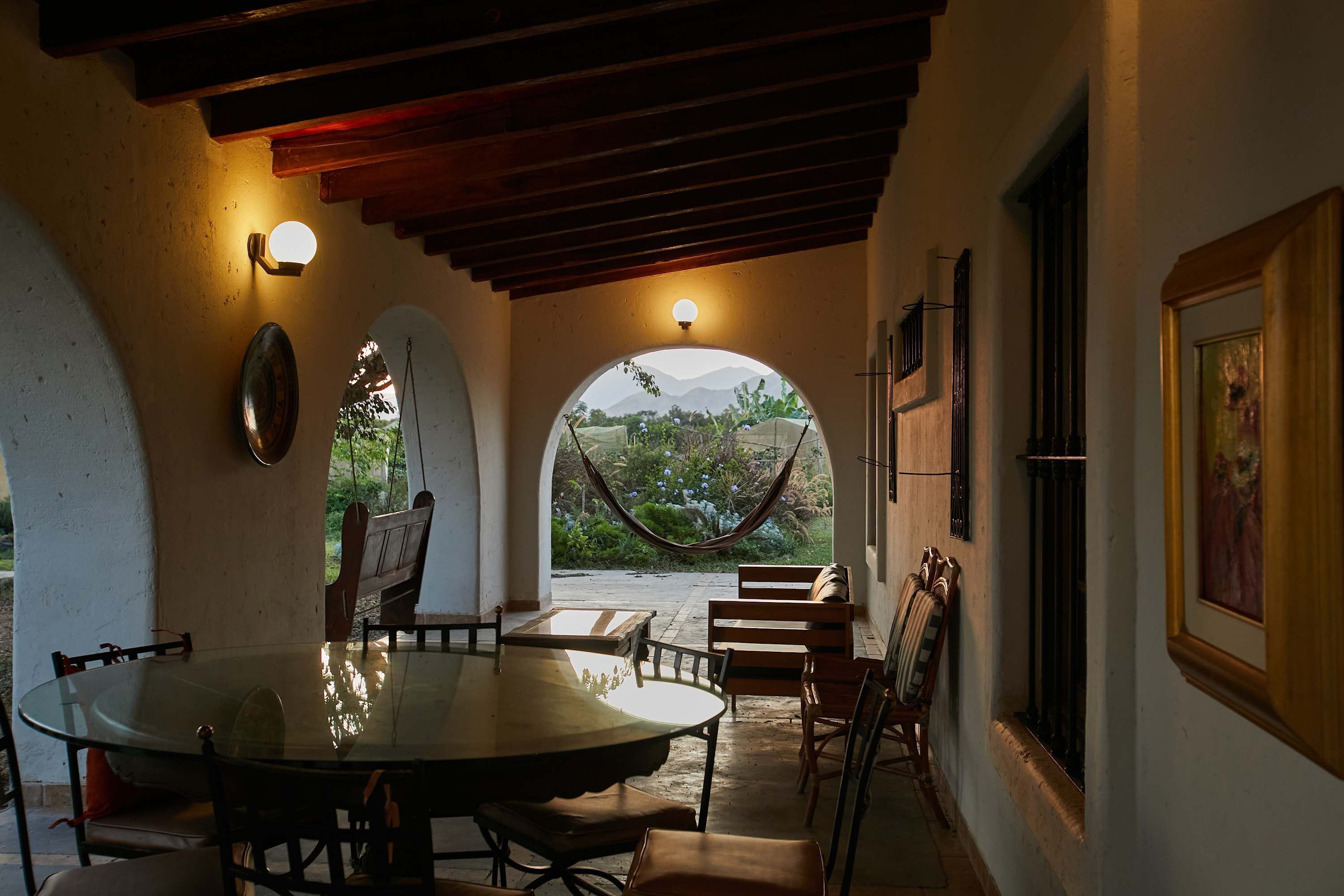 A covered patio with a table and chairs, a hammock, and a view of greenery and mountains through an archway.