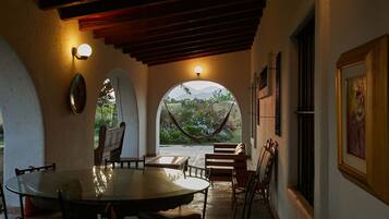 A covered patio with a table and chairs, a hammock, and a view of greenery and mountains through an archway.