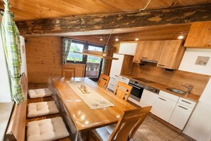 A wooden interior with a dining table, chairs, a kitchen area with cabinets and appliances, and a window with curtains. - Kranvogllehen holiday farm (Schönau am Königssee)