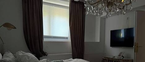 A bedroom with a bed, a television, a chandelier, and a window with curtains.