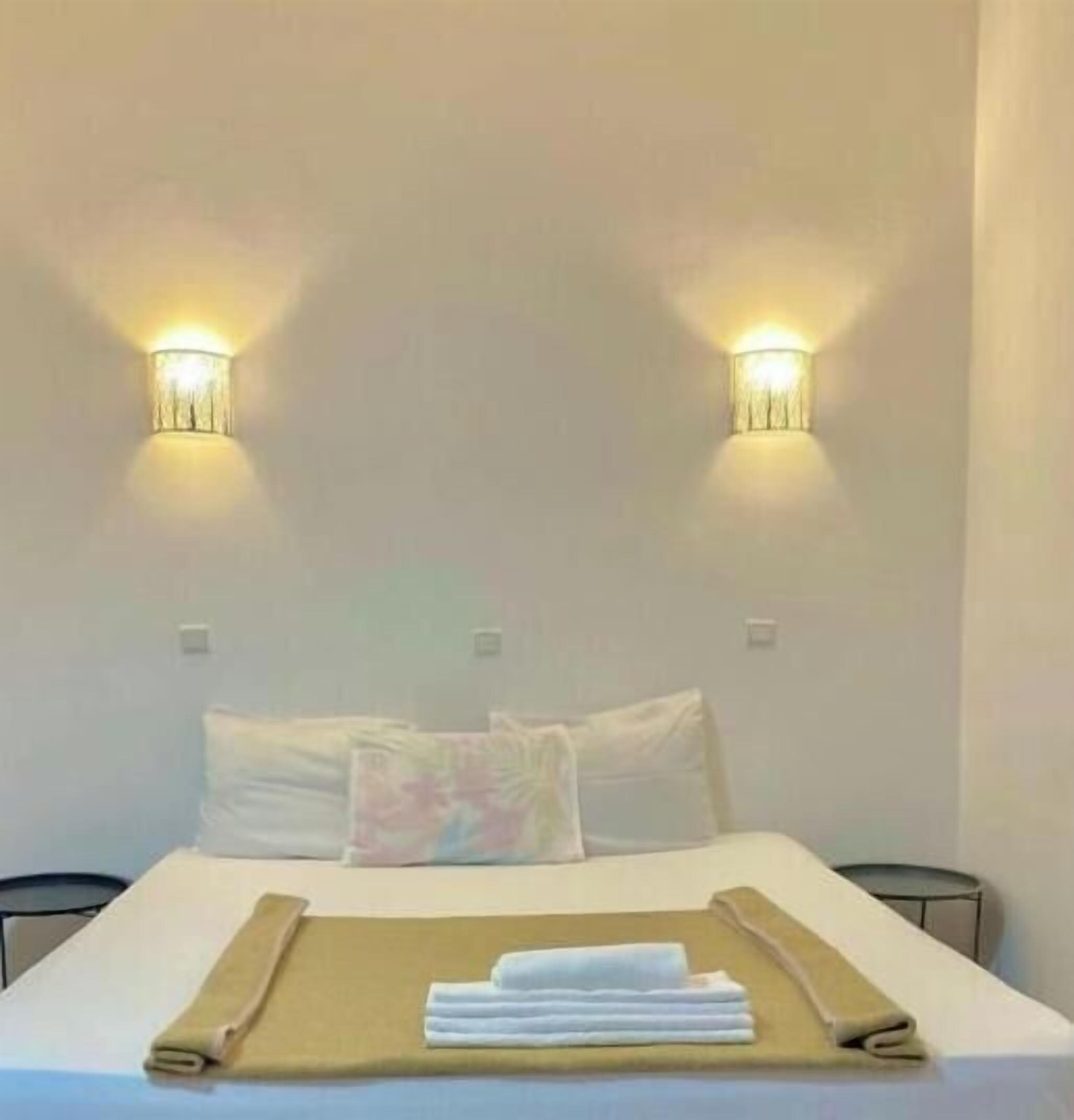 A hotel room with a bed, two wall-mounted lights, a bedside table, and a folded towel on the bed.
