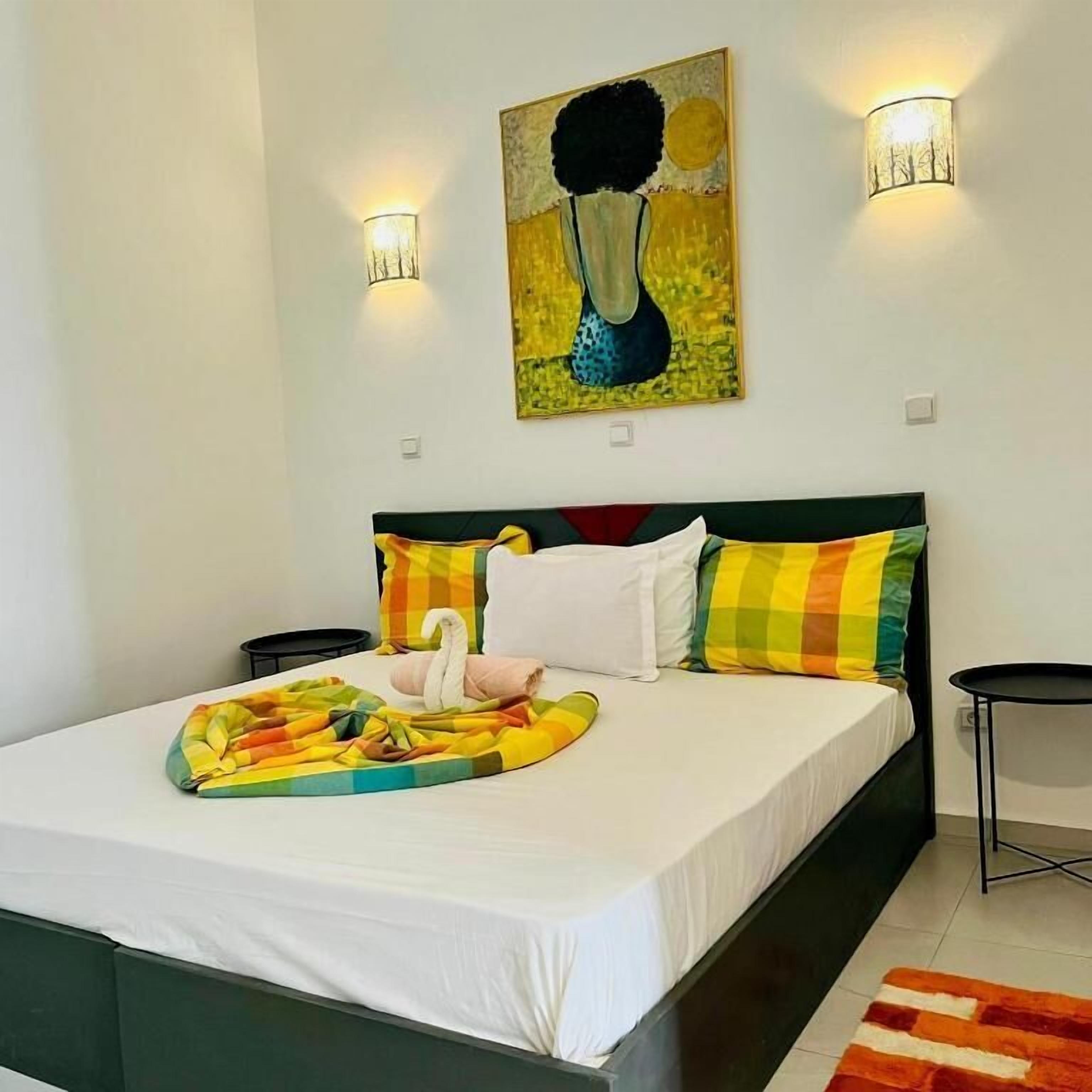 Una cama con colchón blanco, almohadas coloridas, una almohada decorativa con forma de cisne y una manta colorida. Hay dos lámparas de pared y un cuadro sobre la cama.