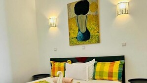 Una cama con colchón blanco, almohadas coloridas, una almohada decorativa con forma de cisne y una manta colorida. Hay dos lámparas de pared y un cuadro sobre la cama.