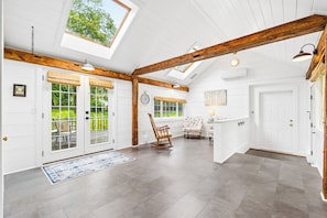 A spacious room with a high ceiling, wooden beams, a skylight, and a view of greenery outside.