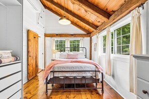 Un dormitorio con techo de madera, cama con estructura metálica, ventana con cortinas y una puerta con una toalla colgada.