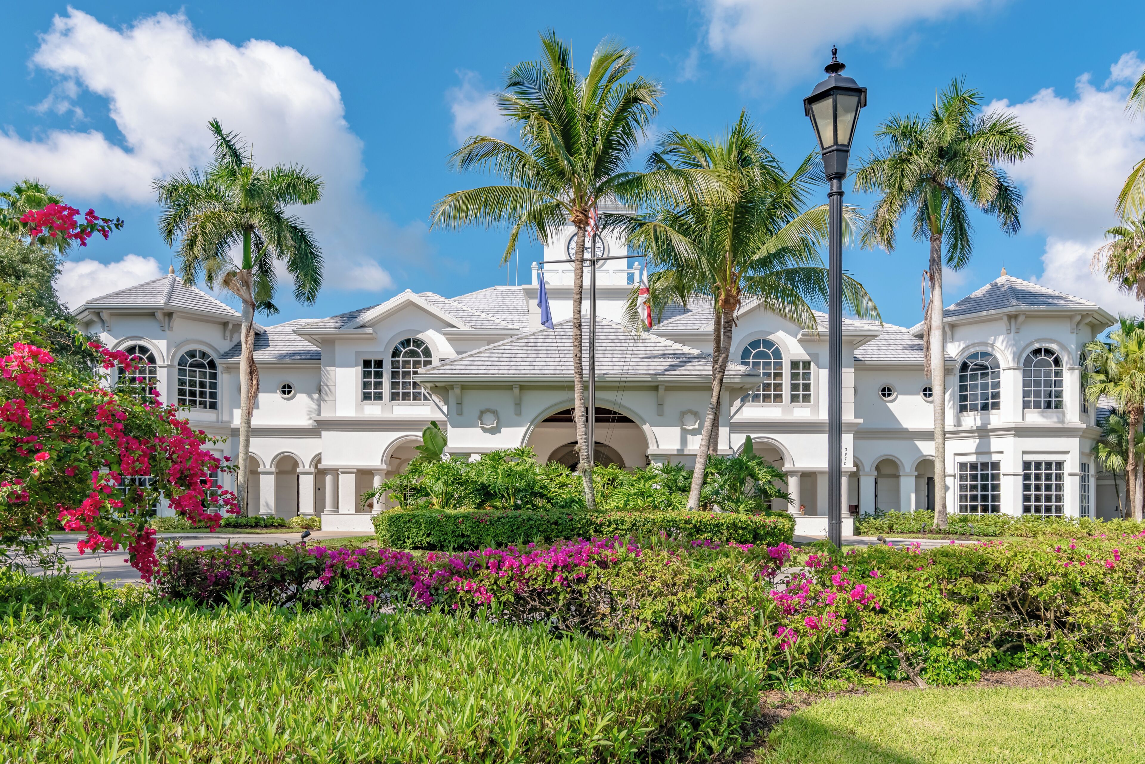 A large, white, two-story building with arched windows and a symmetrical facade, surrounded by lush greenery and palm trees.