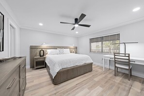 Ein Schlafzimmer mit Bett, Nachttischen, Schreibtisch, Stuhl, Deckenventilator und einem Fenster mit Jalousien.