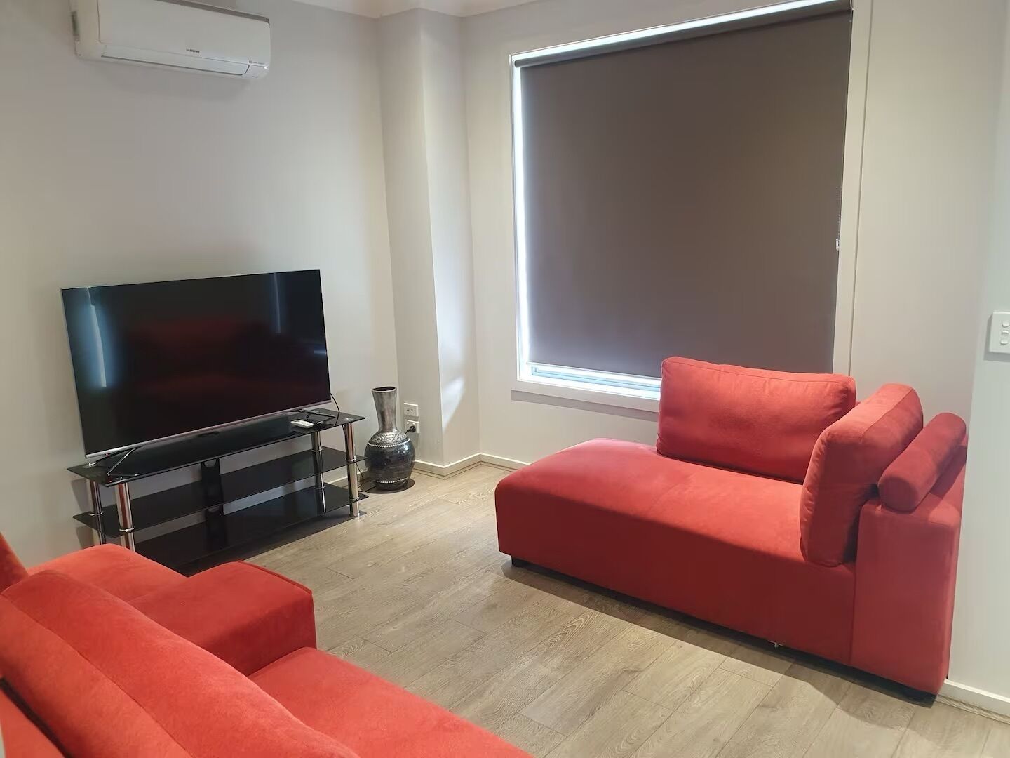 Una sala de estar con una televisión de pantalla plana sobre un mueble bajo, un ventanal con persianas y dos sofás rojos.
