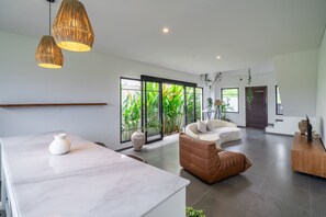 A modern living room with a marble coffee table, a sofa, and a brown leather ottoman. There are large windows with a view of greenery outside. - Senja Villa – Peaceful modern villa in tropical Bali (Kediri)