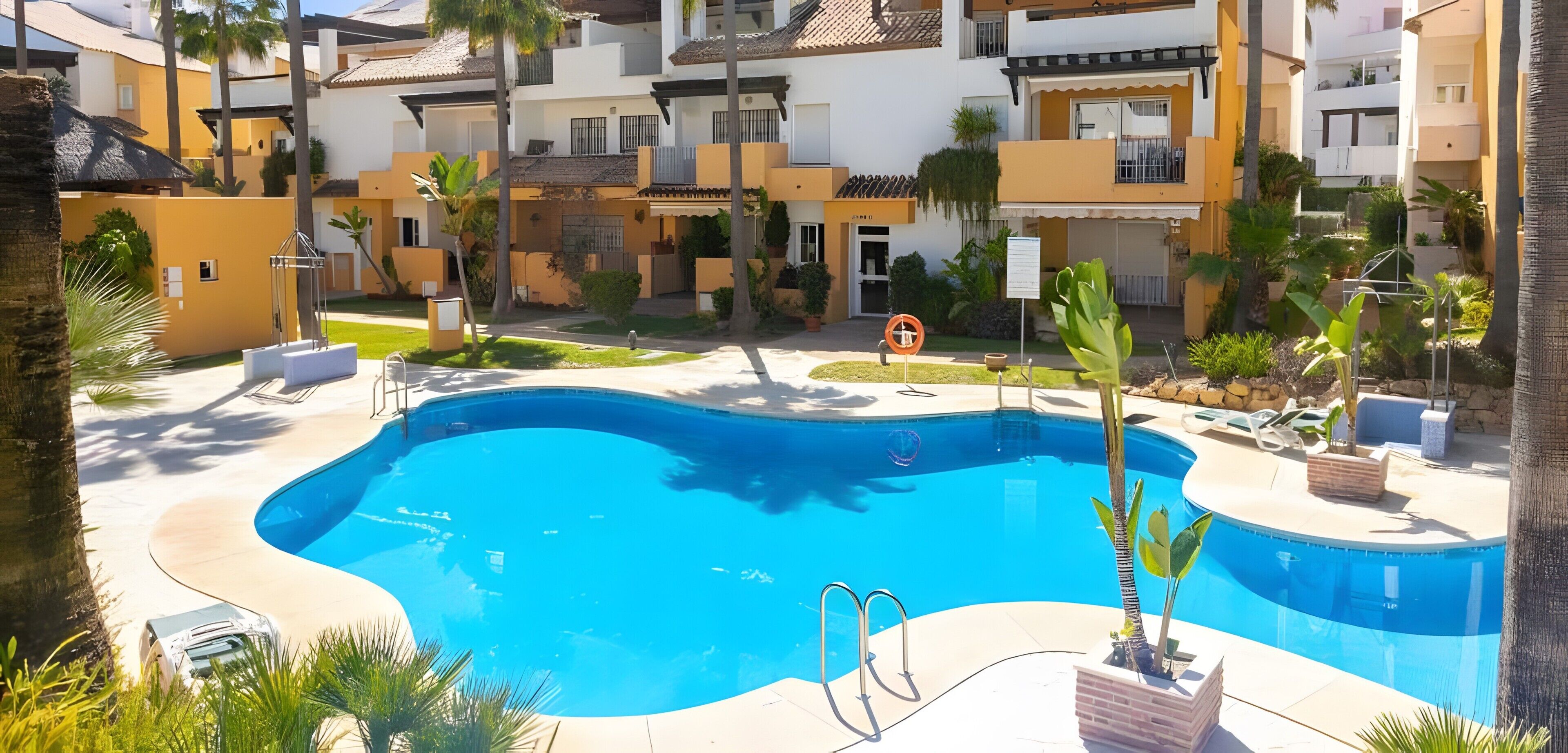 Une piscine entourée d’un espace paysager agrémenté de palmiers, avec un immeuble à plusieurs étages en arrière-plan.