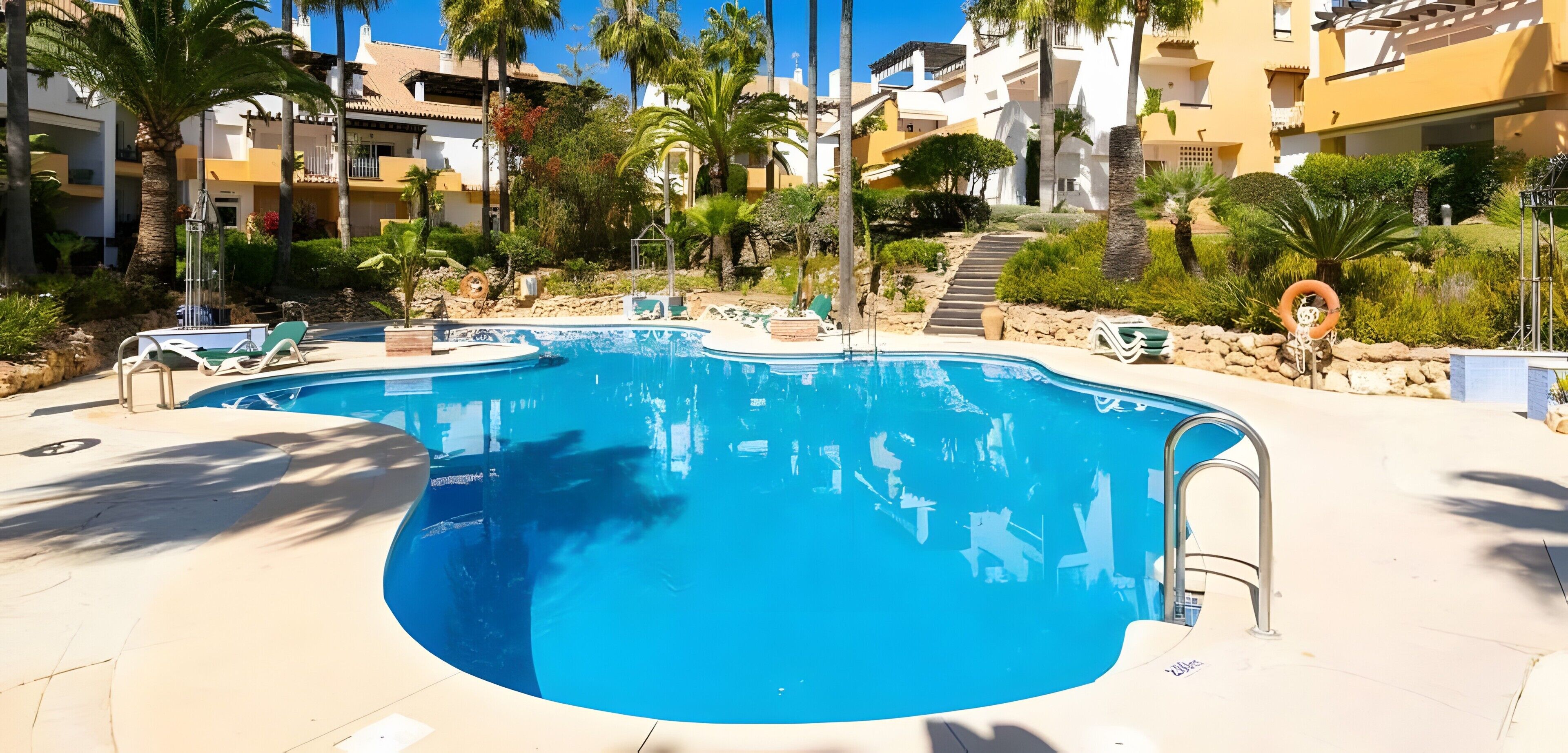 Une piscine dont le centre est plus profond que le reste, entourée de chaises longues et de palmiers.