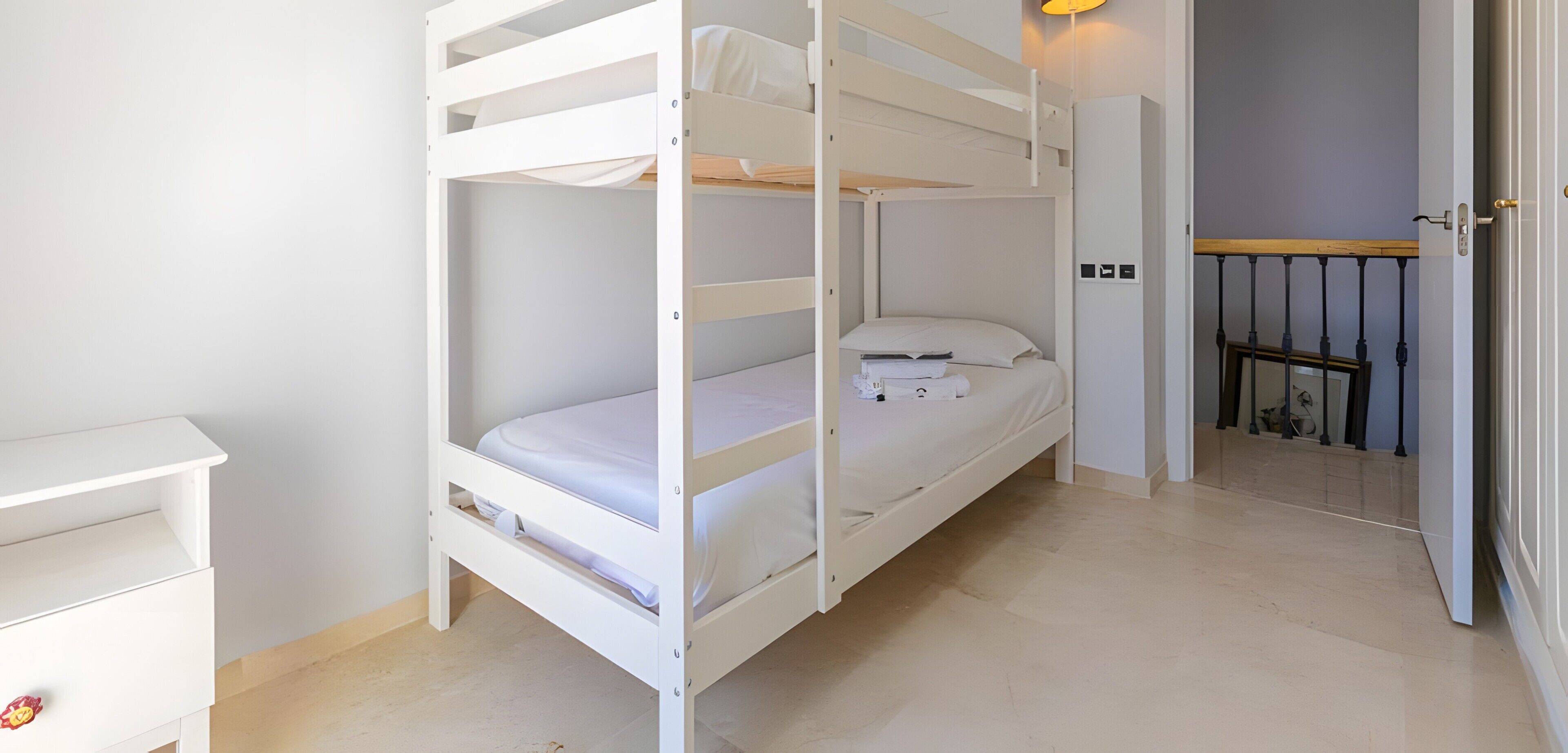 A bunk bed with white bedding, a small bedside table, and a door leading to a balcony with a railing.