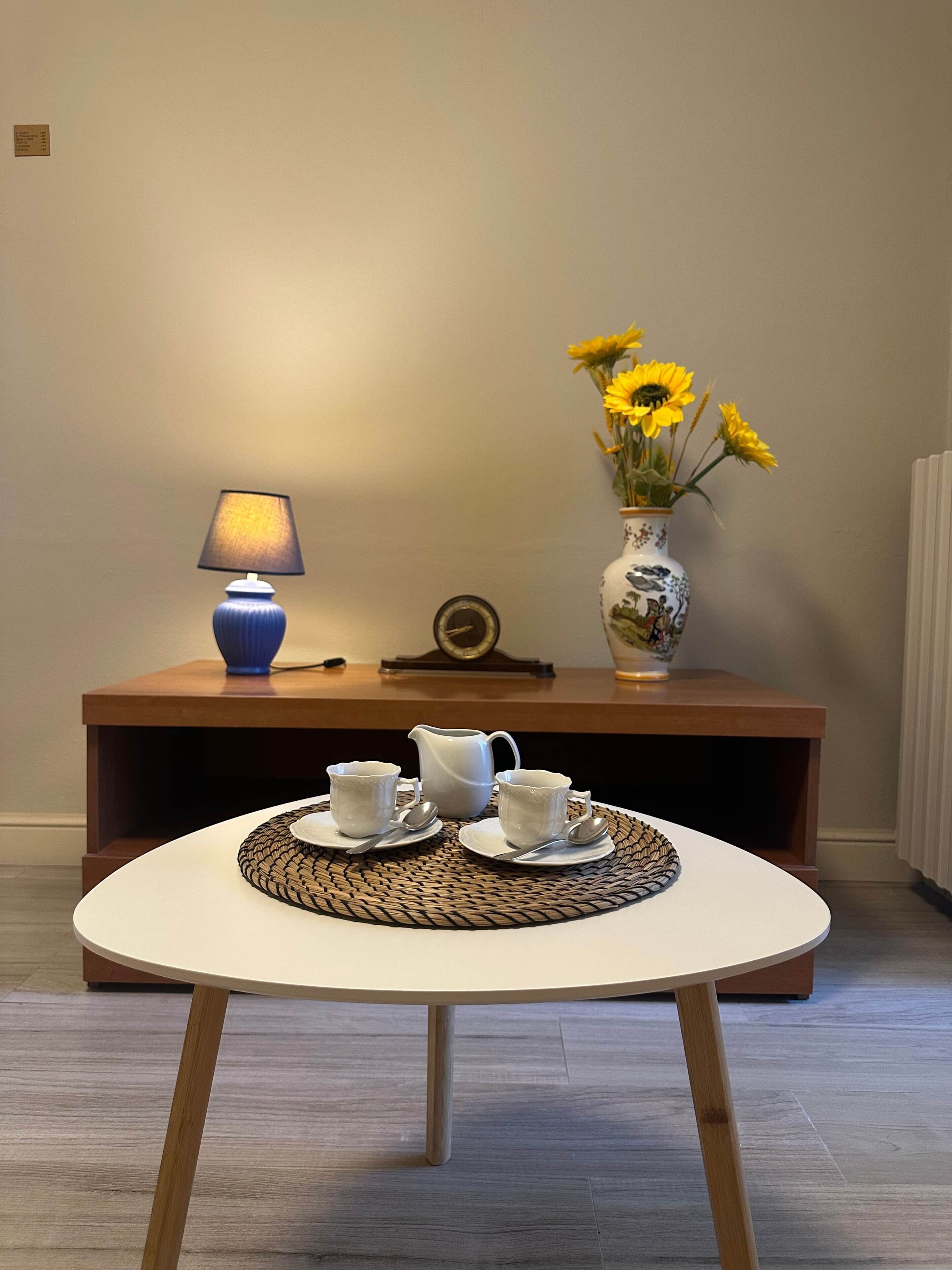 A round table with a woven placemat, two white cups, a white pitcher, and a blue lamp.