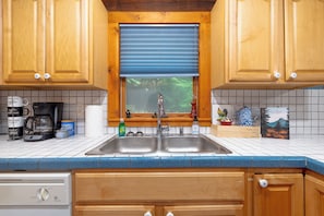 A kitchen with wooden cabinets, a stainless steel sink, a coffee maker, and a window with blinds. - Cozy 2-bedroom cabin in tranquil Sylva with AC (Sylva)