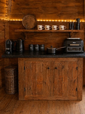 A wooden kitchen with a coffee maker, tea and sugar containers, a Ninja toaster oven, and a shelf with cups.