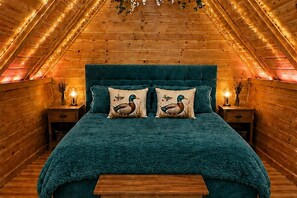 Ein gemütliches Zimmer mit Holzboden und -wänden, einem petrolfarbenen Bett mit passenden Kissen sowie zwei Nachttischen mit Lampen.