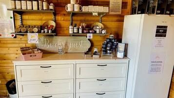 A hotel reception area with a 'Be our guest' sign, a refrigerator with a 'POLITE NOTICE' sign, and various amenities like candles, jars, and a basket of items on shelves.