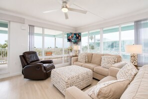 Un salón amplio con un sillón de cuero marrón, sofá beige y reposapiés, ventanas grandes con vistas y un ventilador de techo.