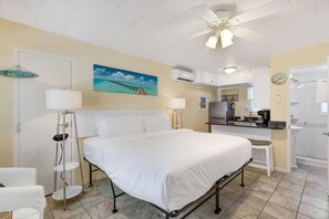 A hotel room with a bed, bedside lamps, a painting of a beach, a ceiling fan, and a kitchenette with a sink and stove. -    🌴Easy Beach Access ☀️ Community Pool/Shuffleboard/Cornhole 🐠 Tropic Terrace #22 (Treasure Island)