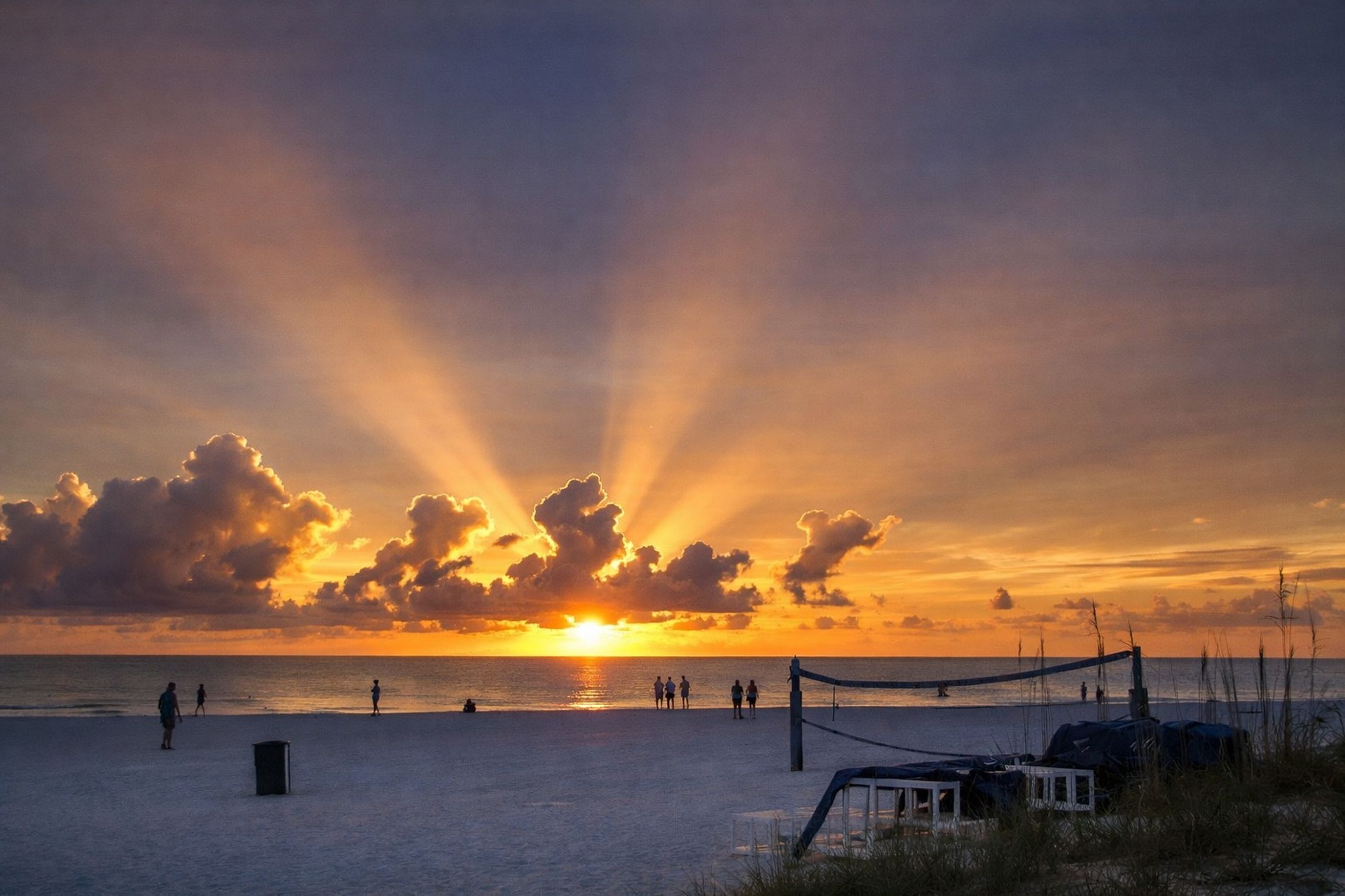 A beach with a volleyball net, people walking, and a sunset.