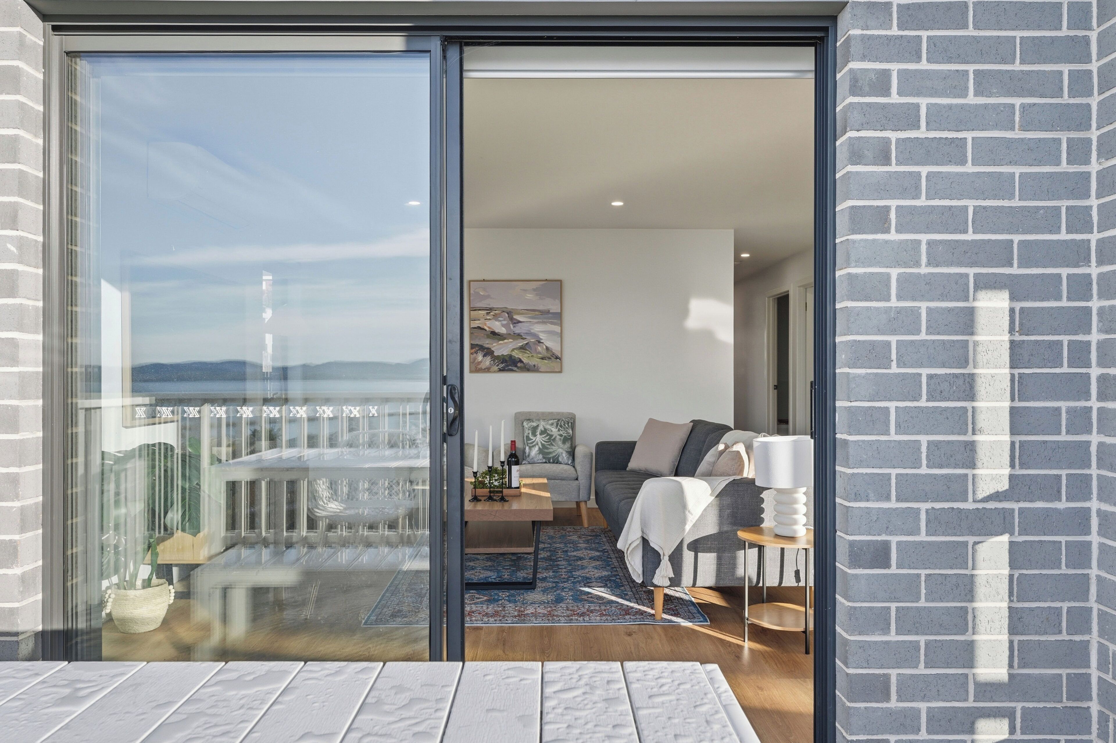 A modern living room with a glass sliding door leading to a balcony, a grey sofa, a coffee table, and a painting on the wall.
