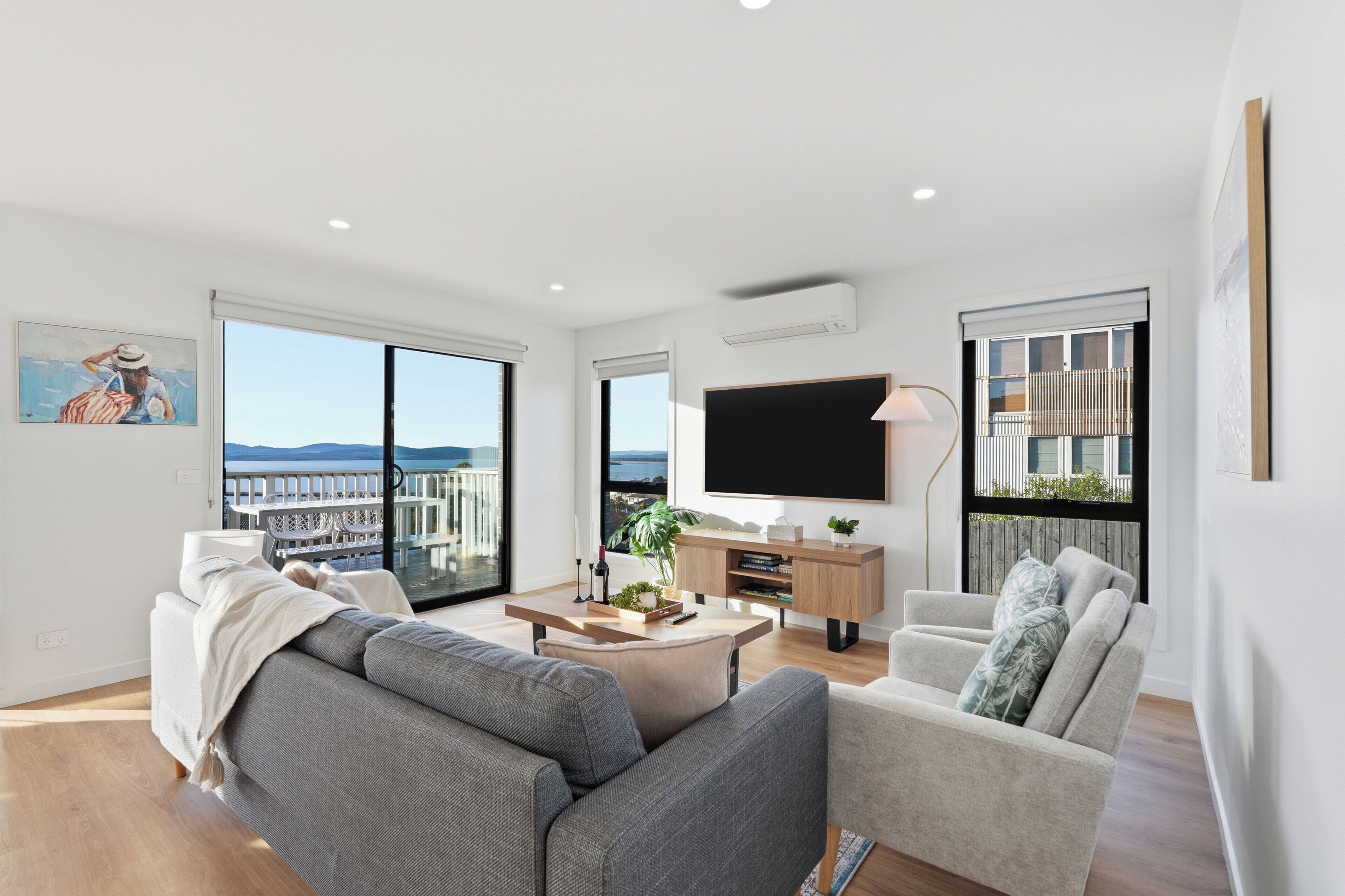 A modern living room with a large TV, comfortable seating, a dining area, and a balcony with a view.