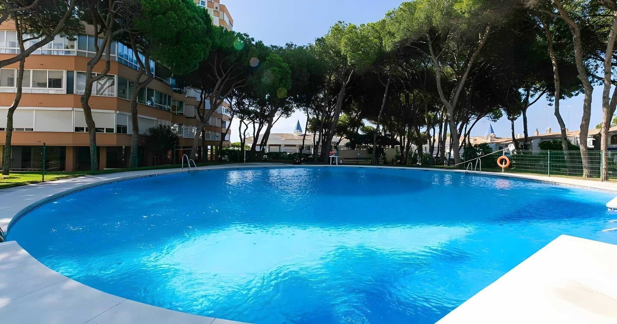 Une piscine à l’eau d’un bleu limpide, entourée d’arbres et bordée d’un bâtiment doté de nombreuses fenêtres.