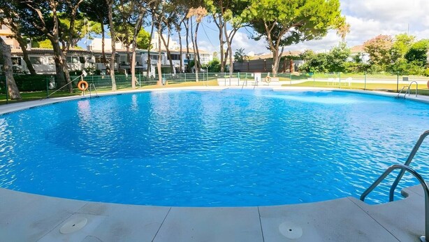 Una piscina azul cristalina con una escalera metálica a la derecha, rodeada por una cerca verde y árboles.