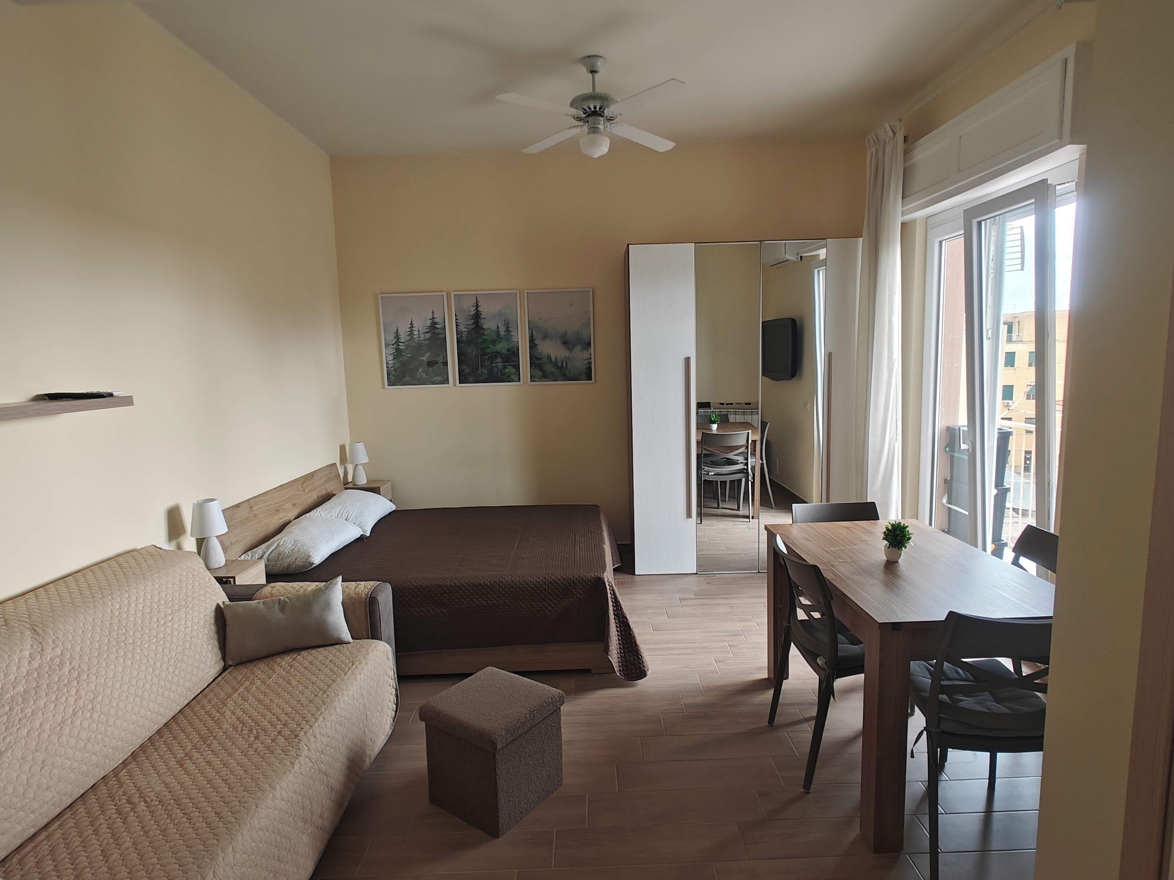 Une chambre à coucher comprenant un lit, un canapé, un bureau et une chaise. On y trouve également un ventilateur de plafond, une fenêtre et une porte donnant sur une salle de bain.