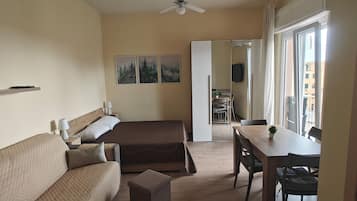 Une chambre à coucher comprenant un lit, un canapé, un bureau et une chaise. On y trouve également un ventilateur de plafond, une fenêtre et une porte donnant sur une salle de bain.