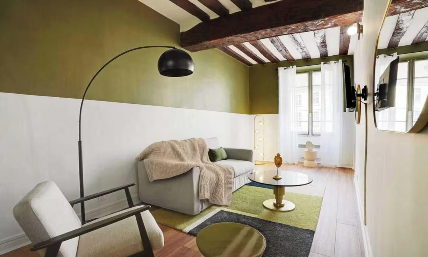 A room with a sofa, a chair, a round table, and a lamp. There is a window with curtains and a view of the outside.