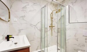 Un baño con ducha envuelta en cristal, un lavabo blanco y paredes con diseño de mármol.