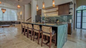 A modern kitchen with a central island, bar stools, and a dining area with a sofa. - 10-bedroom- all Suites - Wellness Compound - Private Sauna/Ice Bath/Yoga Deck (Tulum)