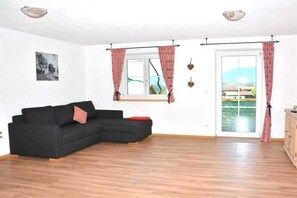 A living room with a black sofa, a wooden floor, a window with pink curtains, and a view of a body of water. - Country House Dream View (Ofterschwang)
